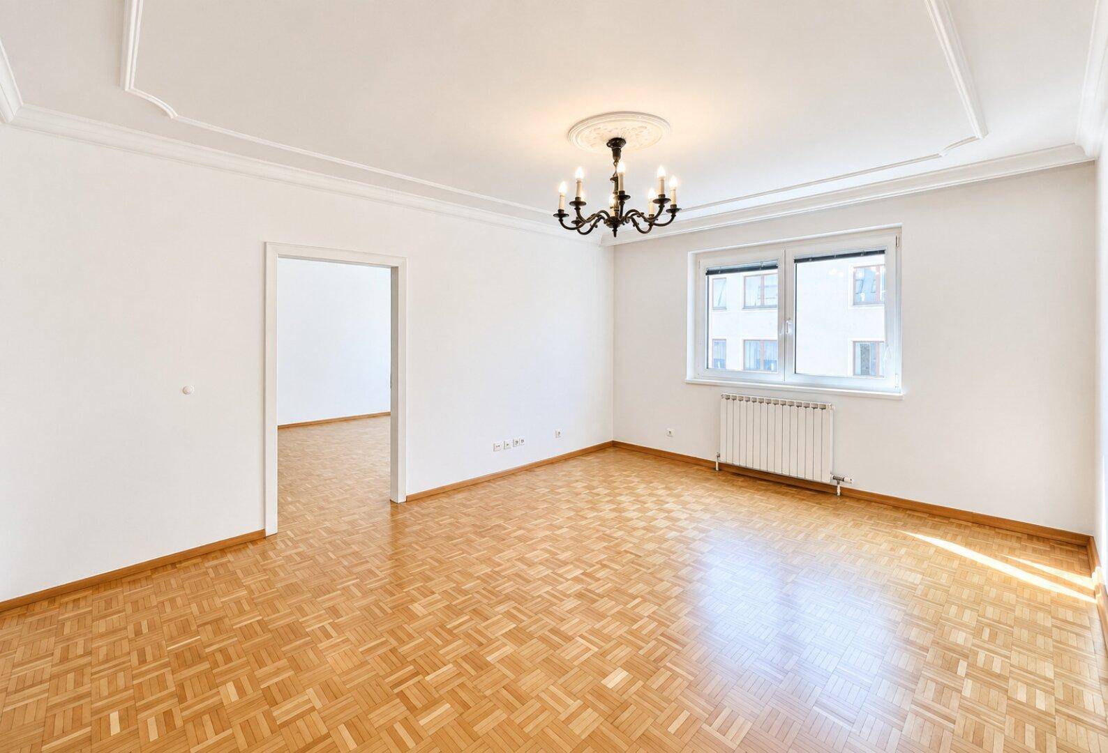Leeres Schlafzimmer mit klassischer Stuckdecke, Parkettboden und viel Tageslicht.