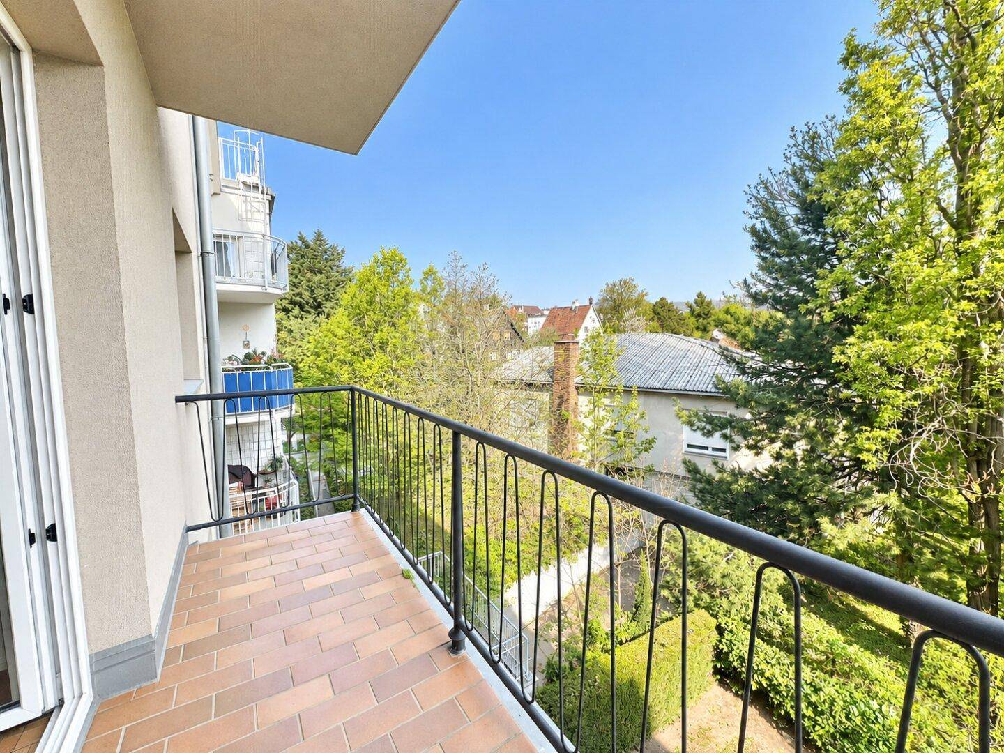 Sonniger Balkon mit Terrakotta-Fliesen und Blick auf den grünen Innenhof und Bäume.