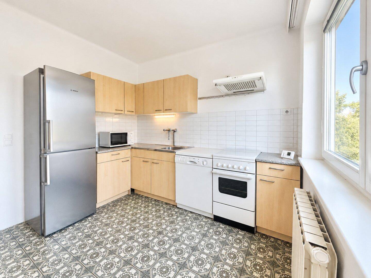 Funktionale Küche mit hellen Holzschränken, Edelstahlkühlschrank und gemusterten Fliesenboden.