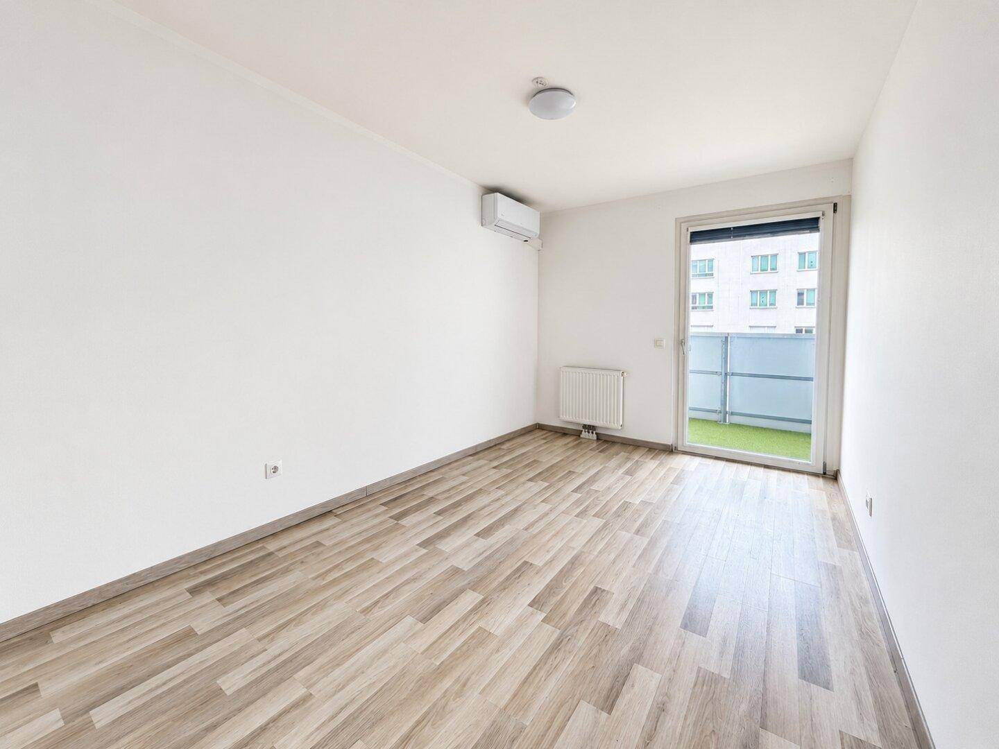 Leeres Schlafzimmer mit Holzboden, weiß gestrichenen Wänden und Zugang zum Balkon.