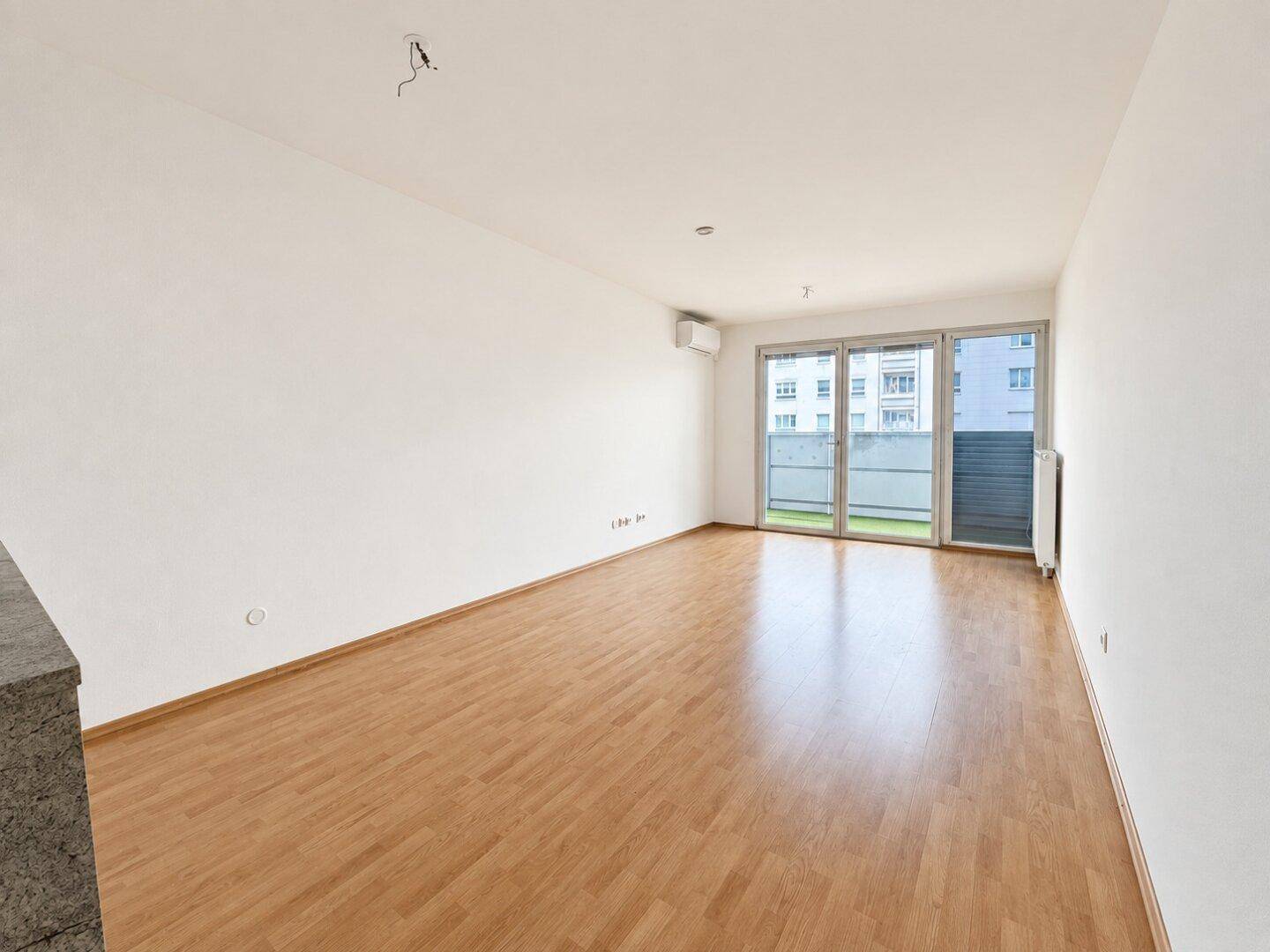 Helles, leeres Wohnzimmer mit Holzboden, großen Fenstern und Zugang zu einem Außenbereich.