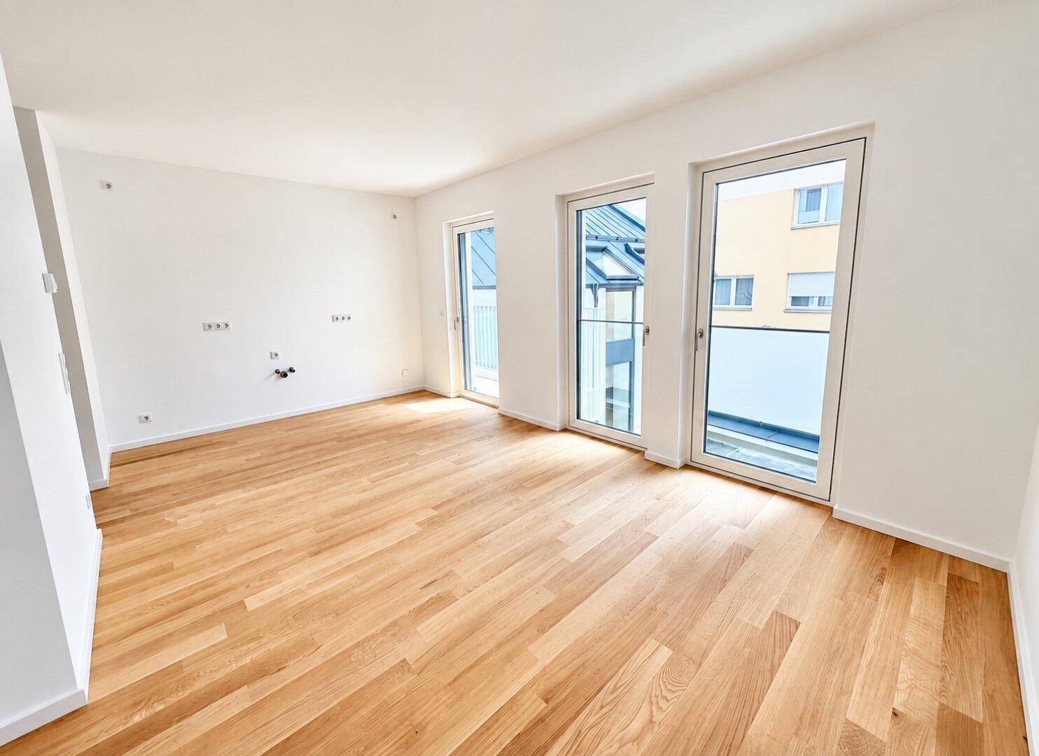 Leerer Küchenbereich mit Eichenparkett, großen Fenstern und Anschlüssen für eine zukünftige Einbauküche.