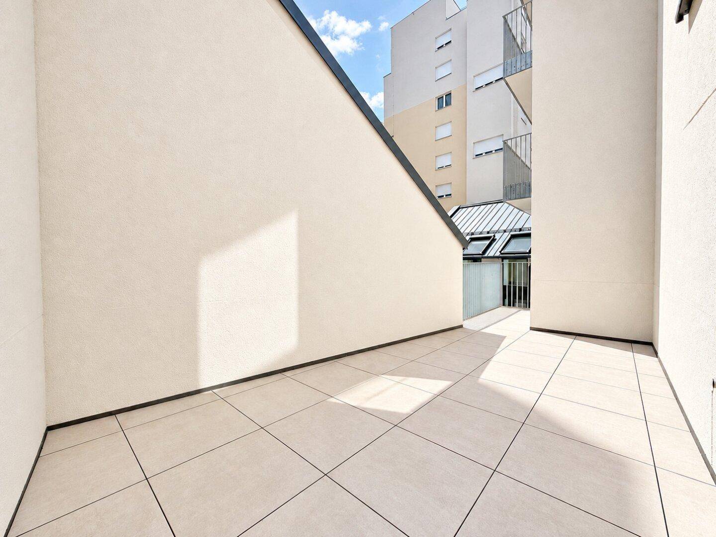 Geräumige, leere Terrasse mit hellen Fliesen und hohen Wänden, umgeben von Gebäuden.