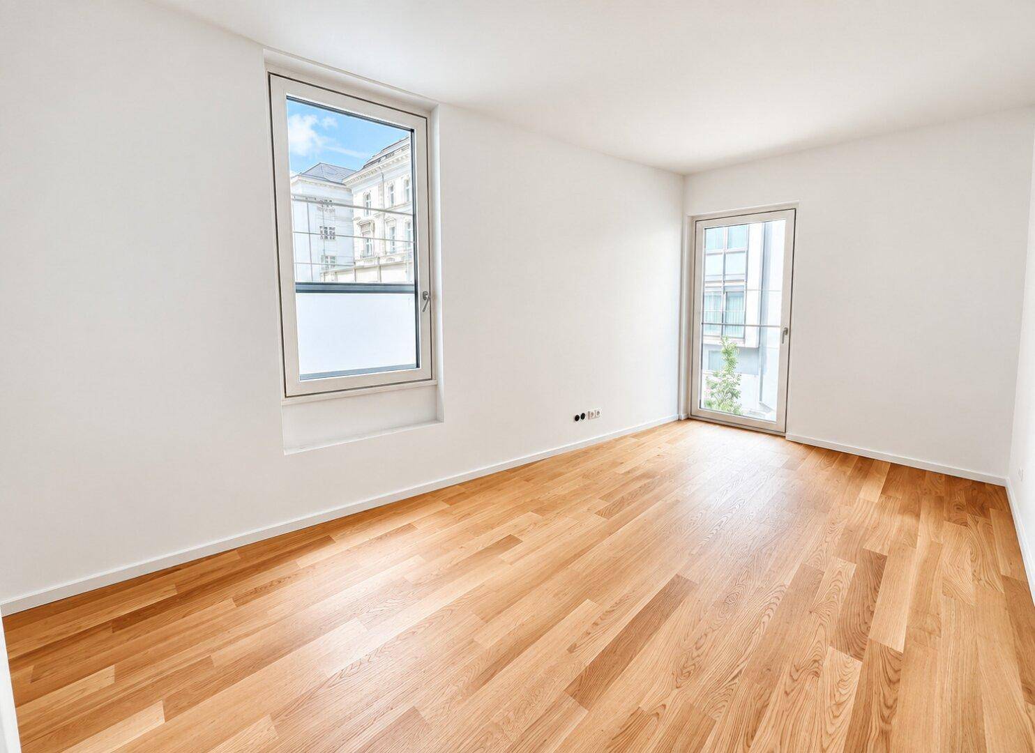 Helles, leeres Schlafzimmer mit Eichenparkettboden, großem Fenster und Zugang zu einem Außenbereich.