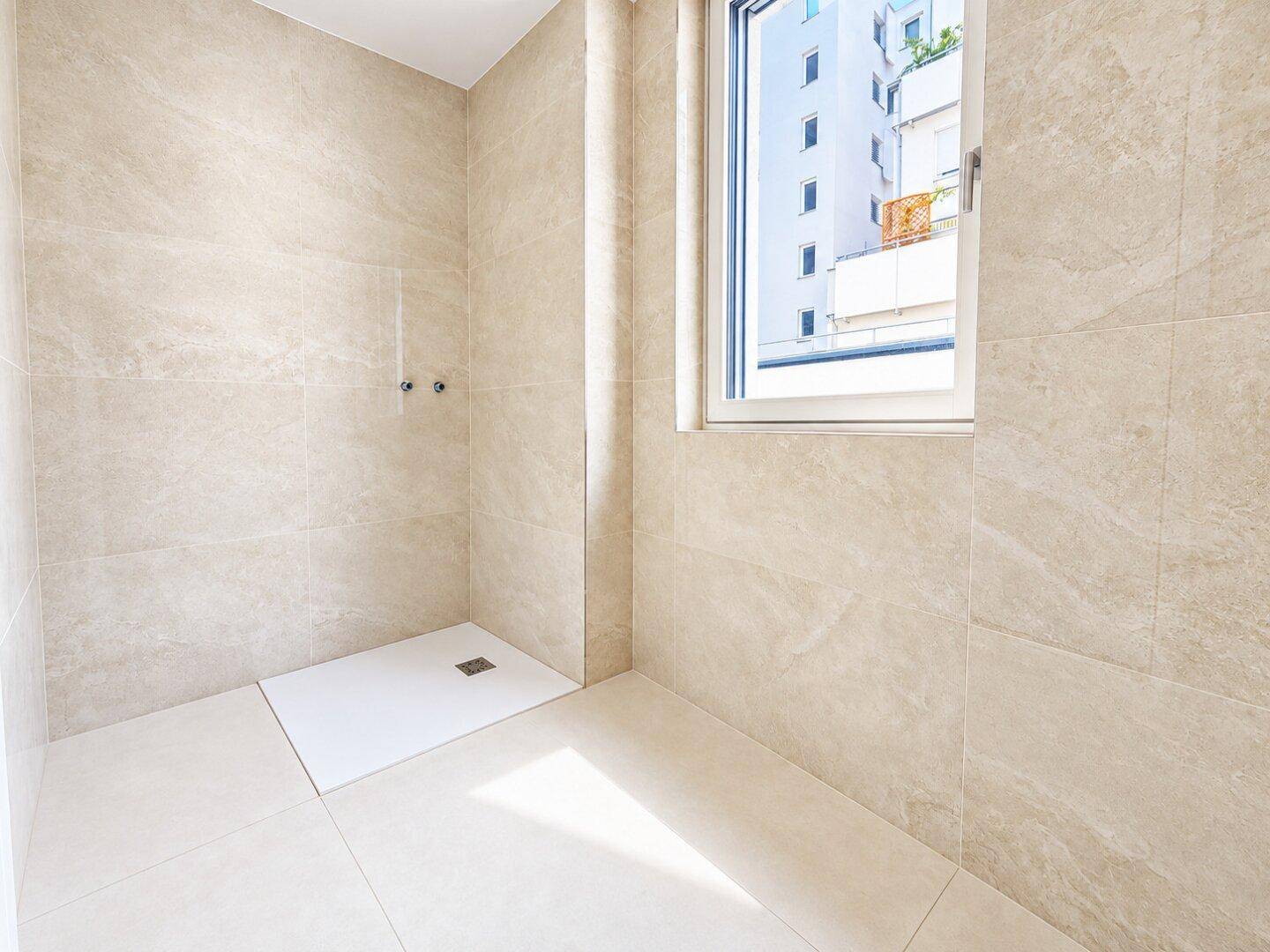 Modernes Badezimmer mit beigefarbenen Fliesen, vorbereitet für eine begehbare Dusche und mit Fenster.