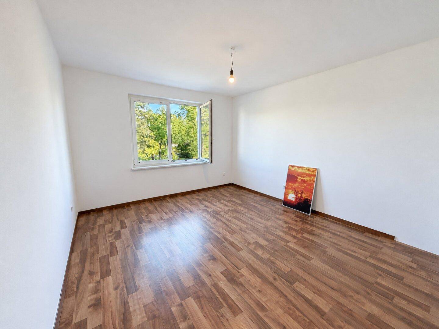 Leeres, helles Zimmer mit Laminatboden und großem Fenster mit Blick ins Grüne.