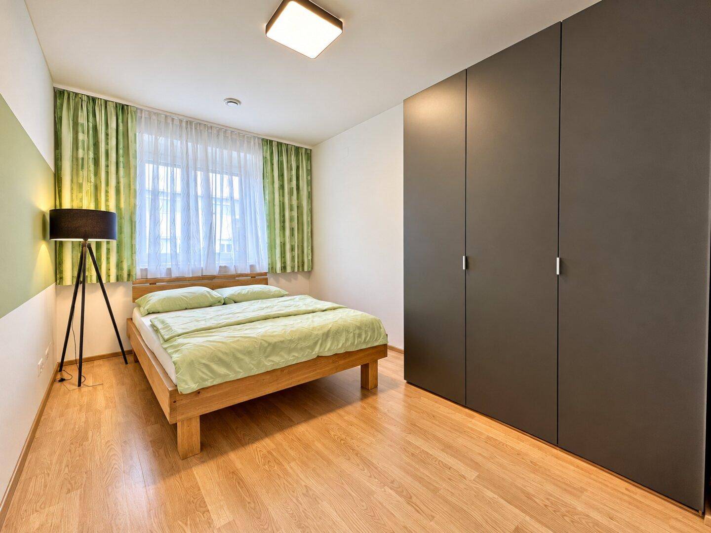 Helles Schlafzimmer mit Holzbett, grünen Vorhängen und großem Kleiderschrank, bietet viel Stauraum.