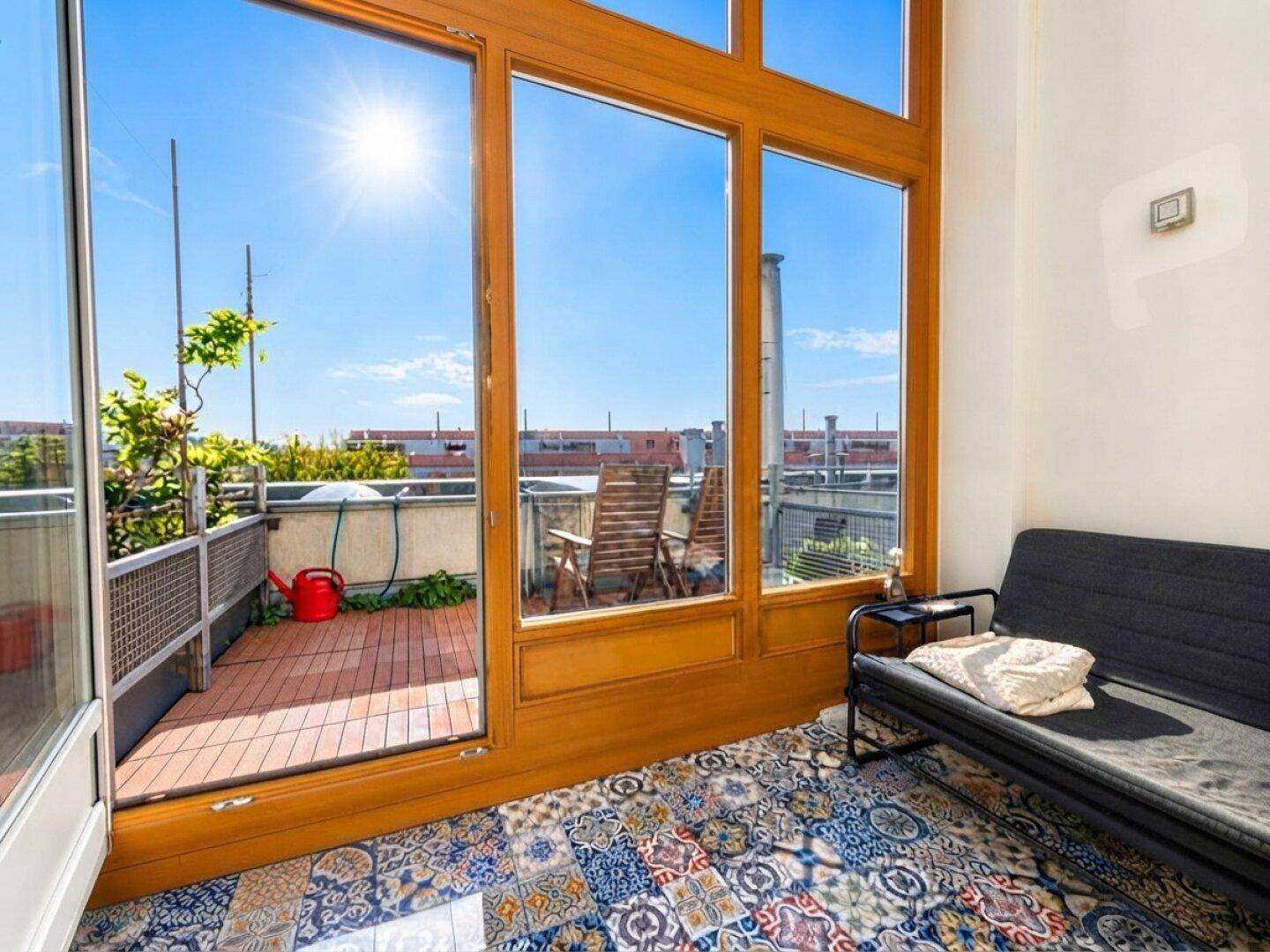 Heller Innenraum mit großen Holzfenstern und Zugang zu einer sonnigen Dachterrasse mit bunten Fliesen.