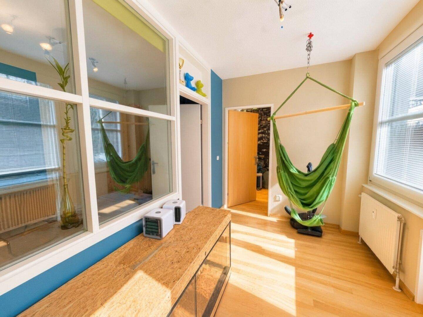 Heller Raum mit Holzfußboden, einer grünen Hängematte und großen Fenstern, ideal zum Entspannen.