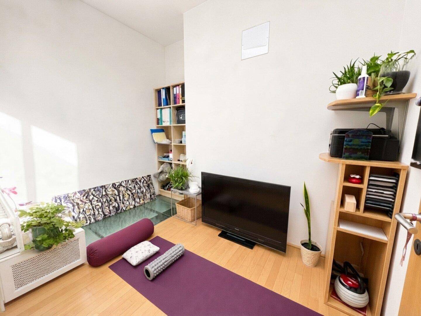 Vielseitiger Raum mit Holzfußboden, Fernseher und Regalen, ideal für Yoga oder entspannte Abende.