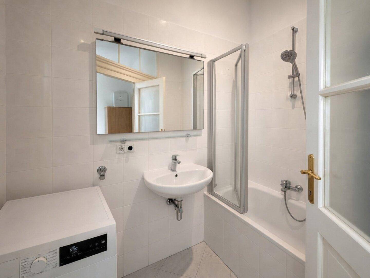 Helles Badezimmer mit weißen Fliesen, Badewanne mit Duschwand und Waschmaschine.
