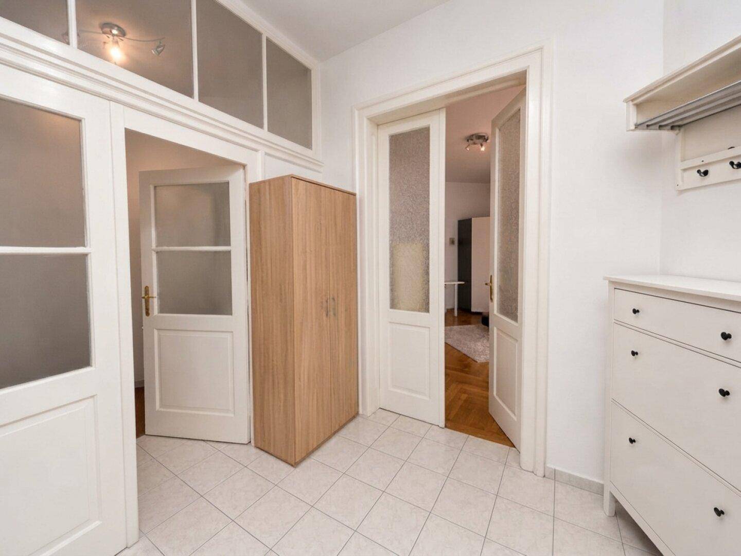 Heller Flur mit weißen Fliesen, Holzkleiderschrank und Türen mit Milchglas.