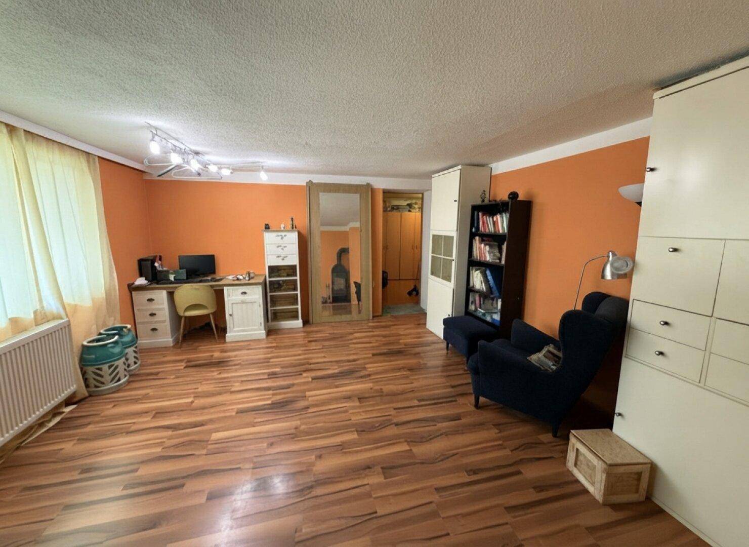 Geräumiges Wohnzimmer mit warmen orangefarbenen Wänden, Laminatboden und einem Arbeitsbereich.