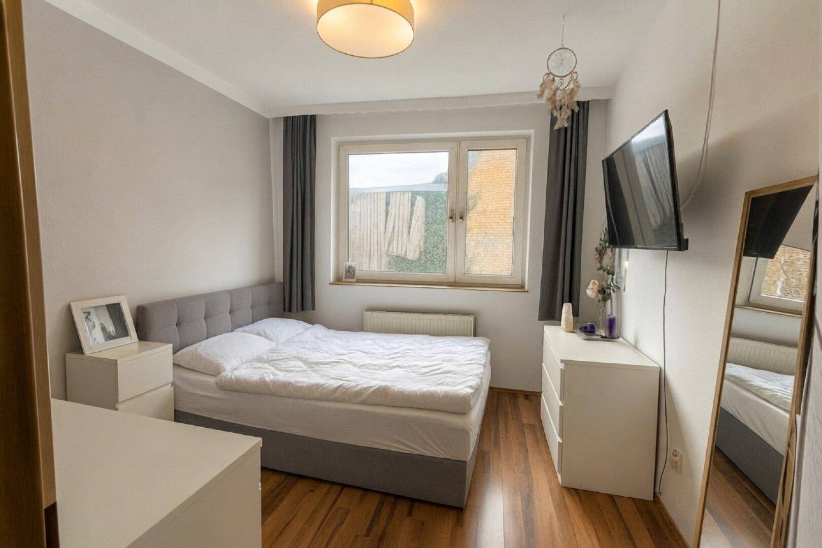 Helles Schlafzimmer mit Doppelbett, Laminatboden und einem Fenster mit Blick ins Grüne.