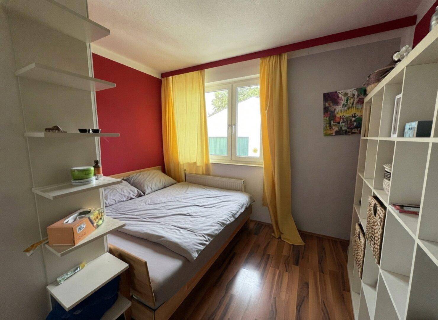 Farbiges Schlafzimmer mit roter Akzentwand, Holzboden und einem Fenster mit gelben Vorhängen.