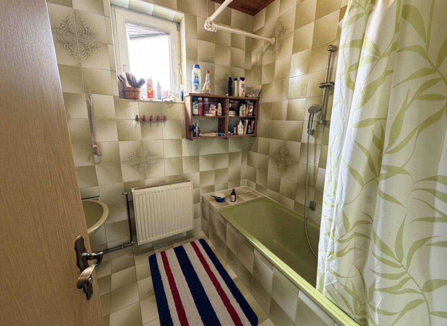 Badezimmer im Stil der 80er Jahre mit grüner Badewanne, gemusterten Fliesen und Fenster.