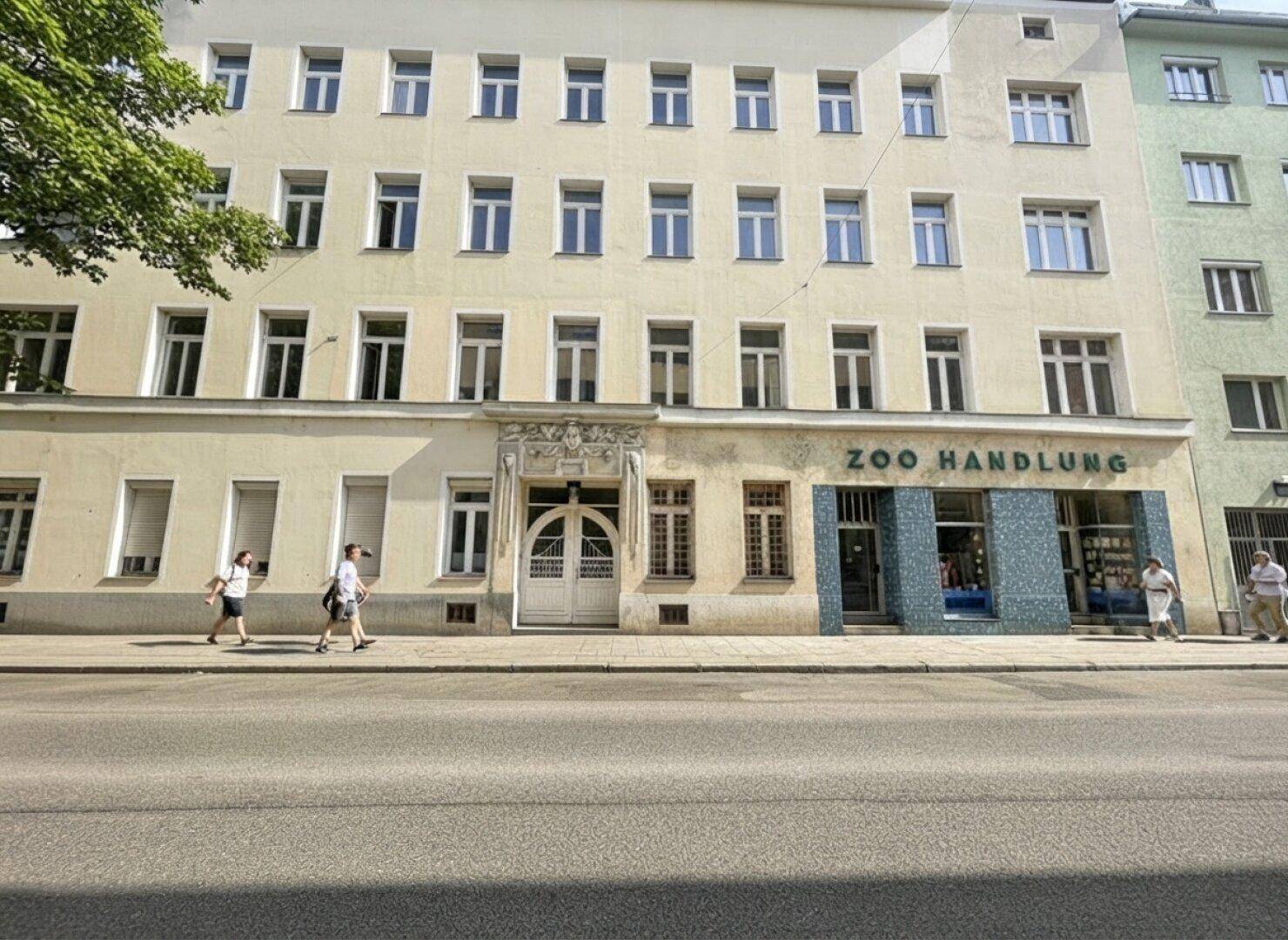 Klassisches Mehrfamilienhaus mit beiger Fassade und Geschäften im Erdgeschoss an einer Stadtstraße.