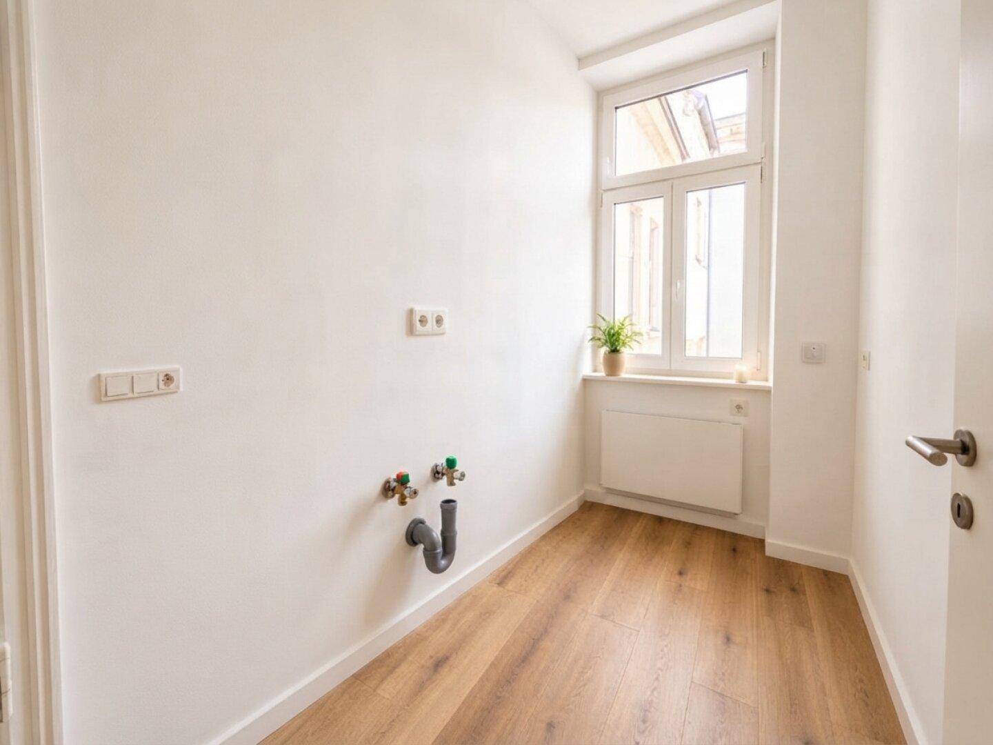 Leerer Raum mit Anschlüssen für eine Küche, Holzboden und einem großen Fenster.
