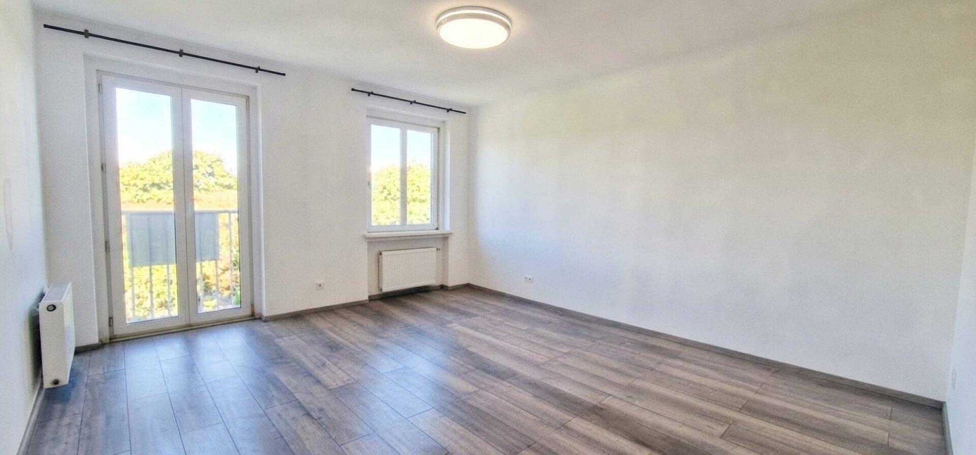 Leerer Raum mit Laminatboden, großen Fenstern und Zugang zu einem französischen Balkon.