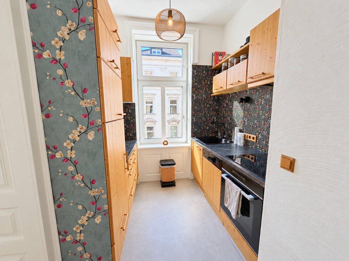 Schmale Küche mit Holzschränken, schwarzer Arbeitsplatte, integriertem Backofen und Fenster.