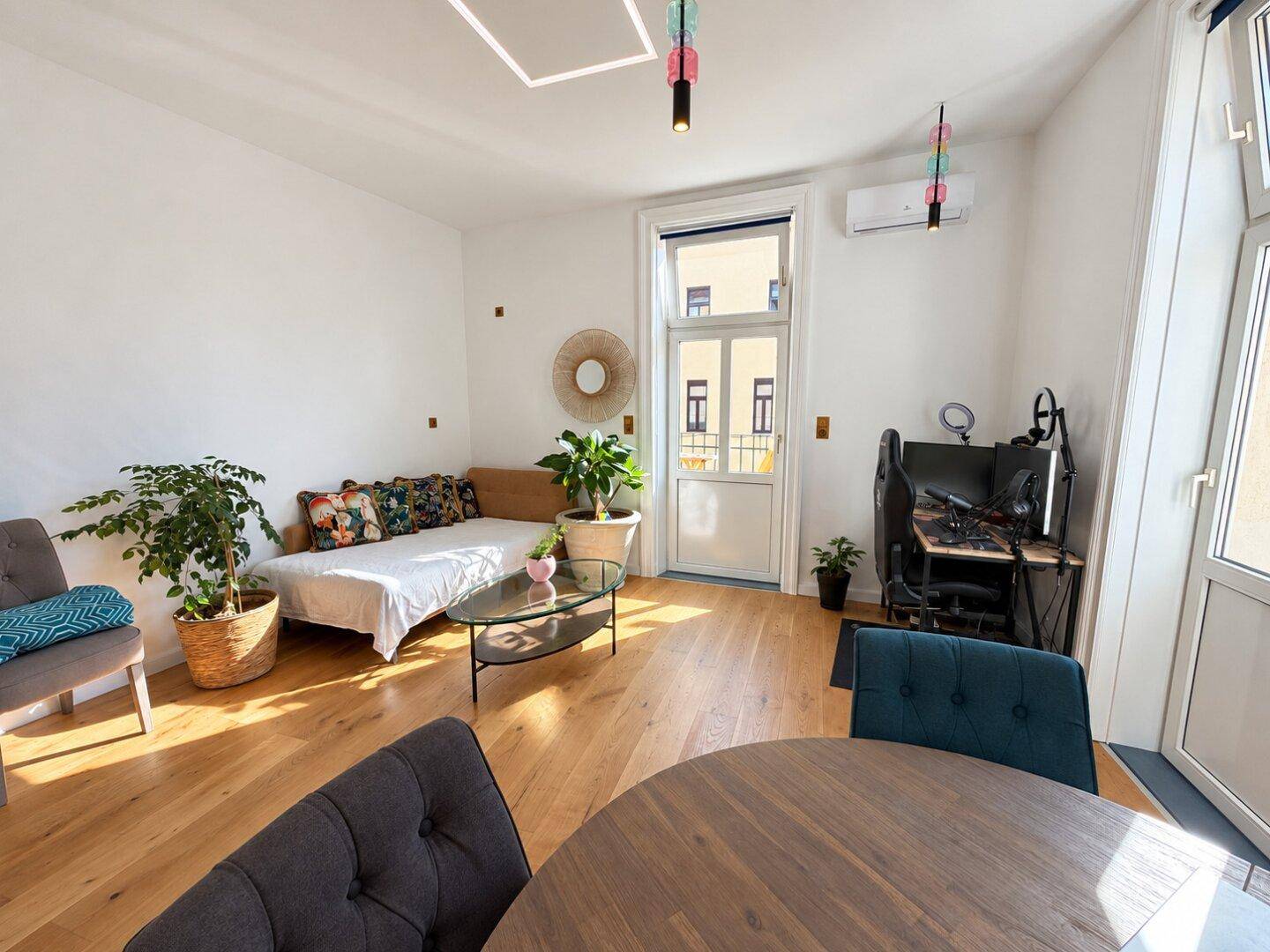 Helles Wohnzimmer mit Holzboden, bequemer Couch, Arbeitsbereich und Zugang zum Balkon.