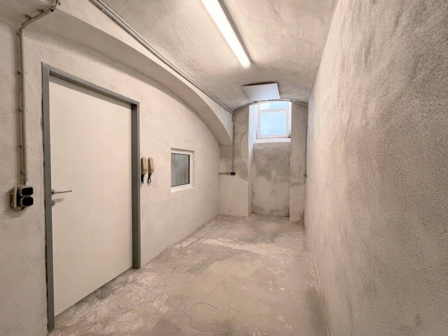 Langer Kellerflur mit Betonwänden, einer Tür und einem kleinen Fenster.