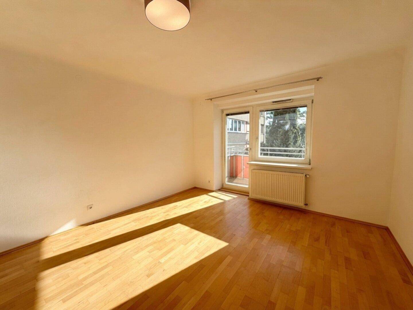Helles Zimmer mit Parkettboden, weißen Wänden und Zugang zu einem Balkon, durchflutet von Sonnenlicht.