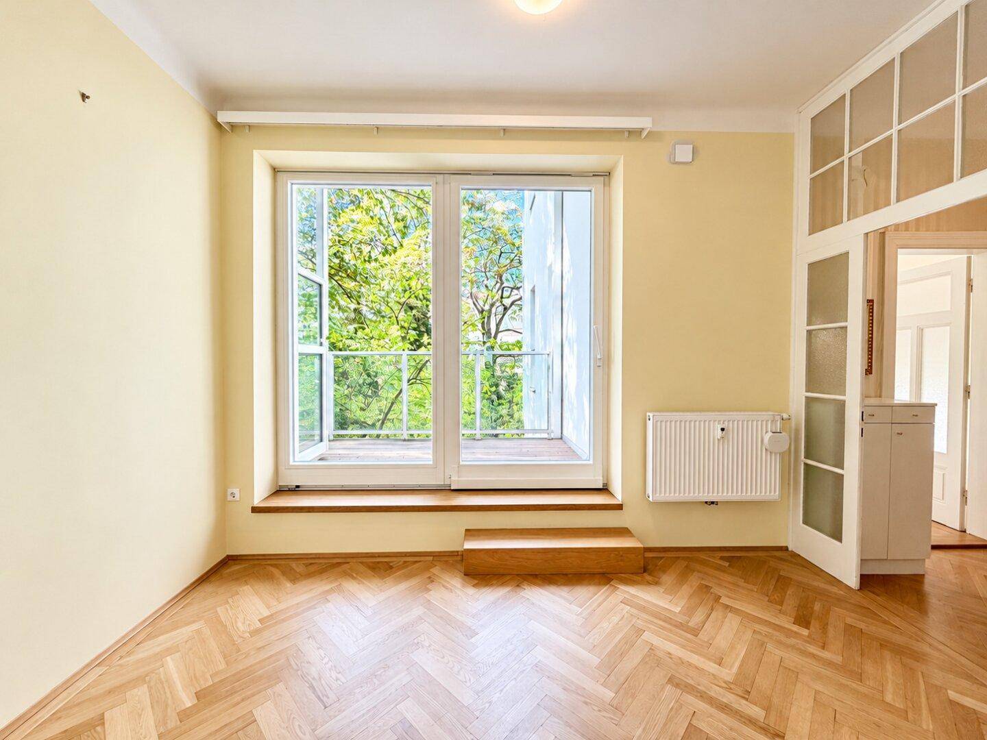 Heller Raum mit Fischgrätparkett und Zugang zu einem französischen Balkon, bietet viel Tageslicht.