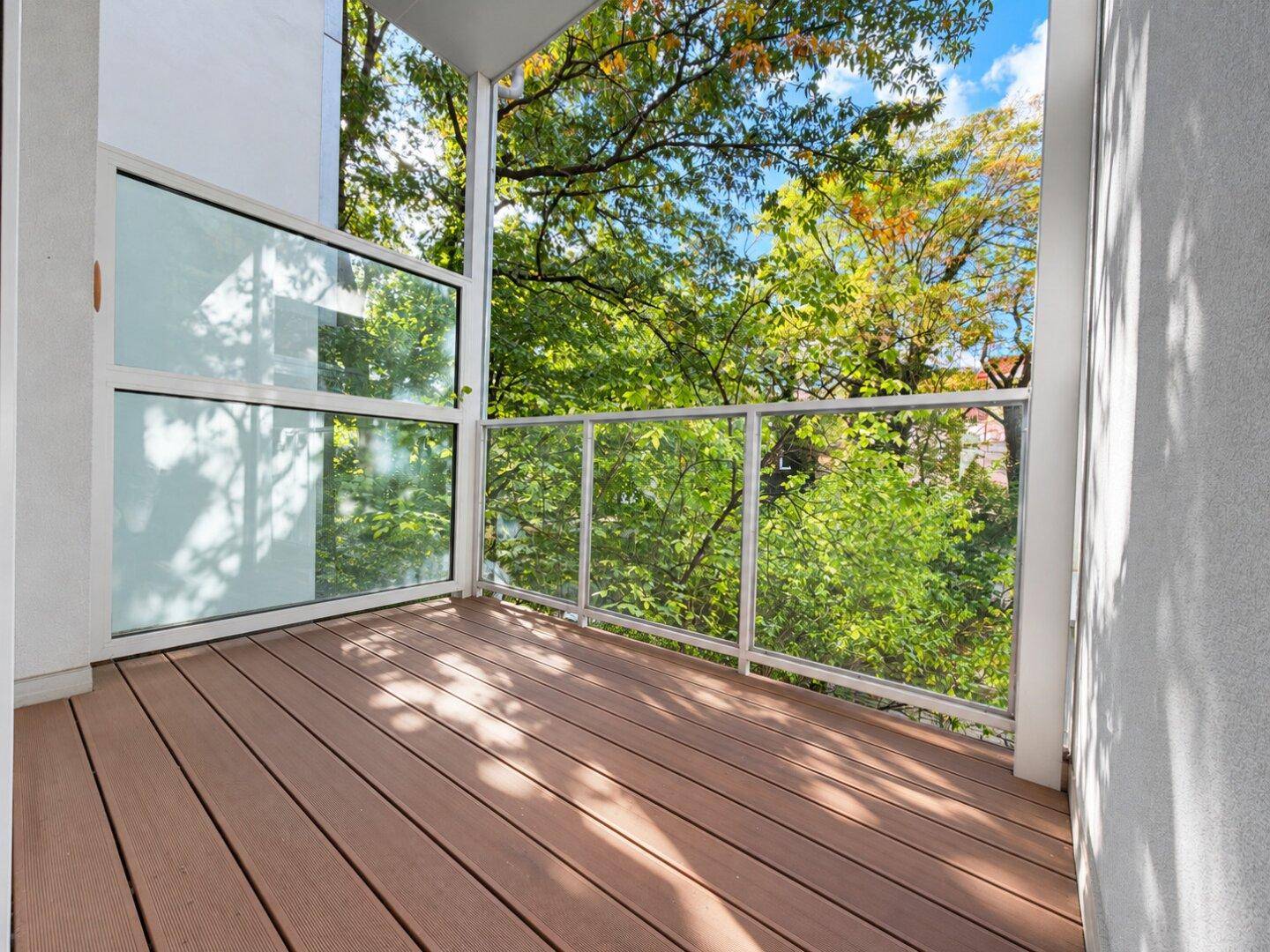 Moderner Balkon mit Holzdeck und Glasgeländer, umgeben von üppigem Grün und Bäumen.