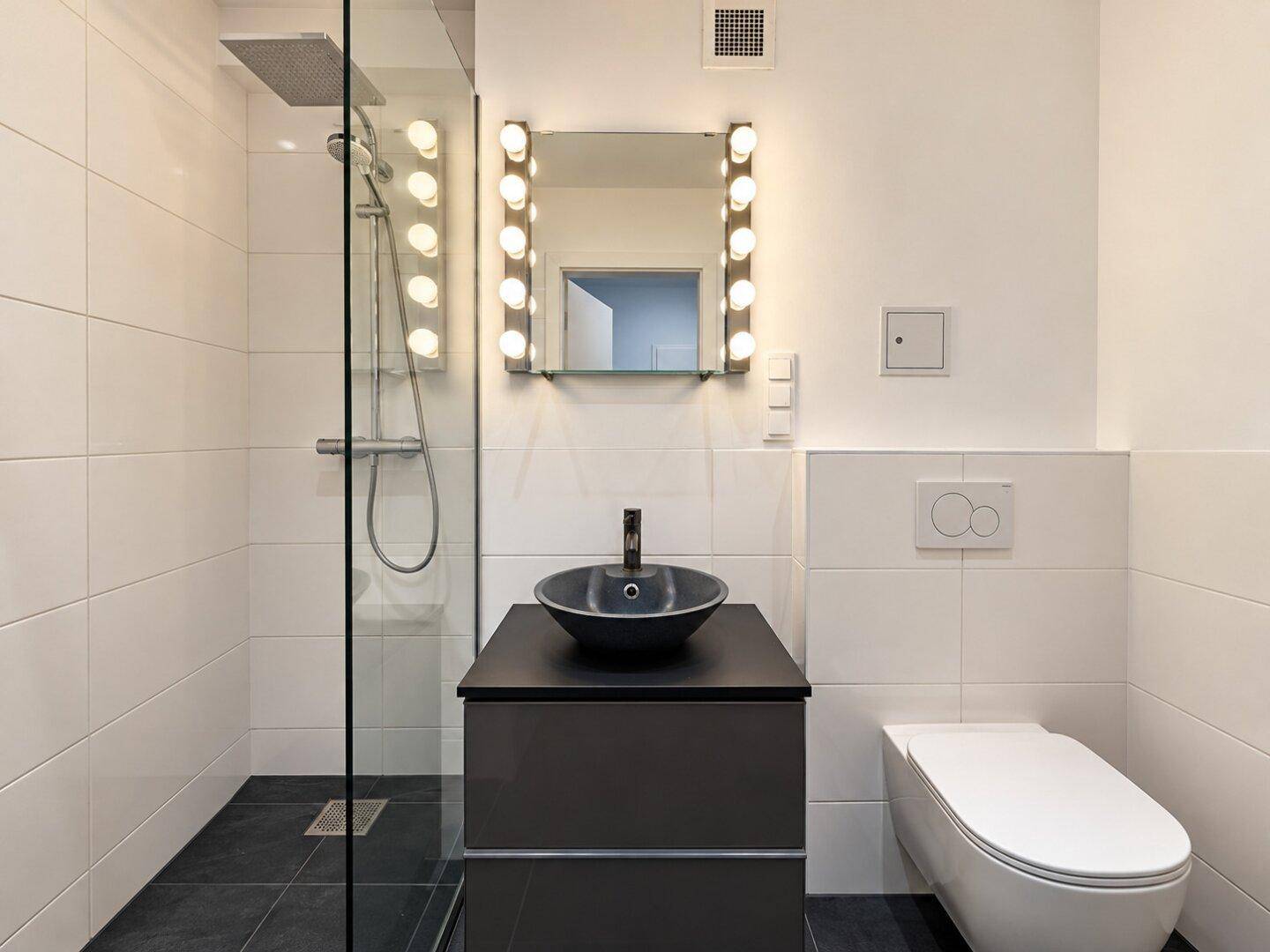 Modernes Badezimmer mit ebenerdiger Dusche, dunklen Fliesen und beleuchtetem Spiegel.