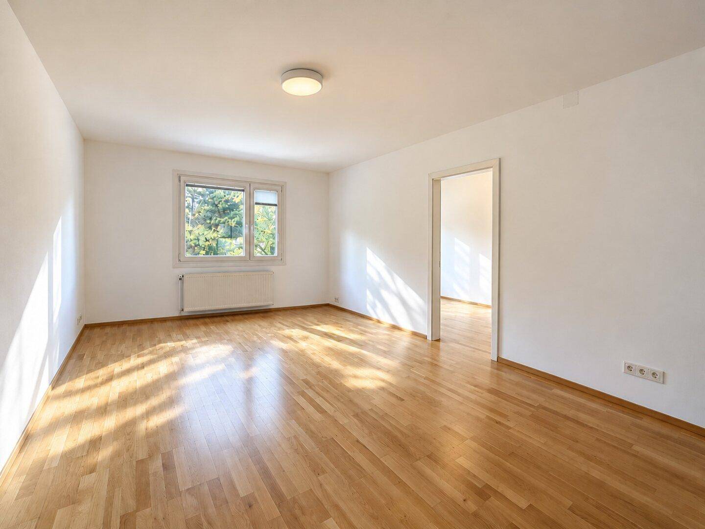 Lichtdurchfluteter Raum mit Holzparkett und Fenster, bietet vielseitige Nutzungsmöglichkeiten und einen Durchgang.