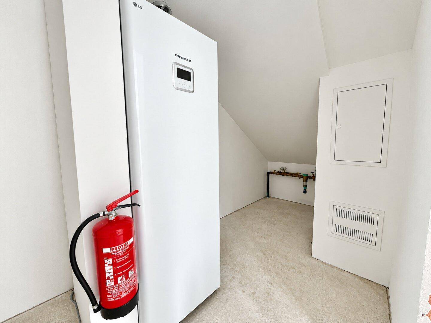 Technikraum mit moderner Wärmepumpe, Feuerlöscher und elektrischer Verteilung unter Dachschräge.