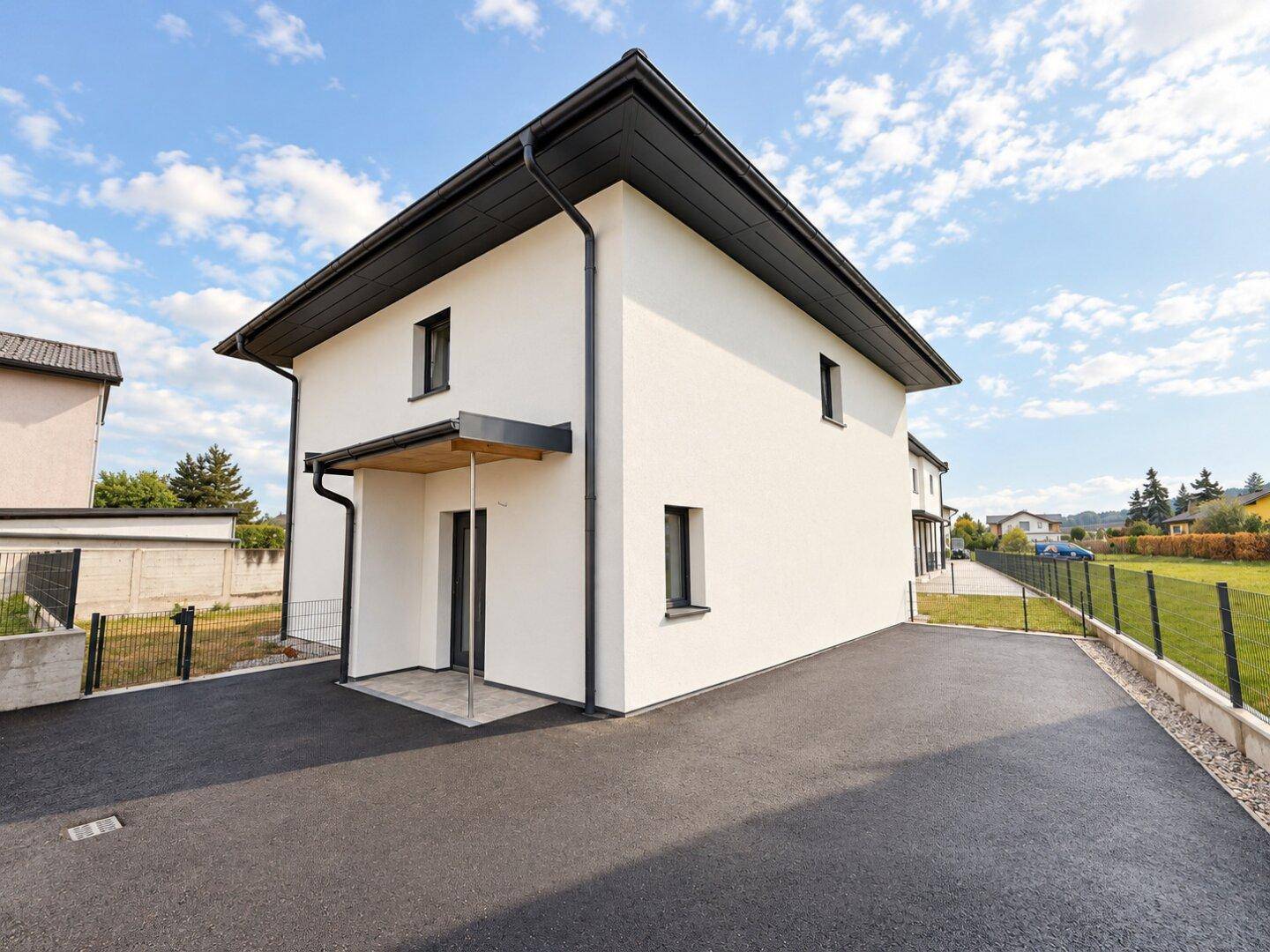 Modernes Einfamilienhaus mit weißer Fassade, dunklem Dach und asphaltierter Zufahrt, umgeben von Grünflächen.