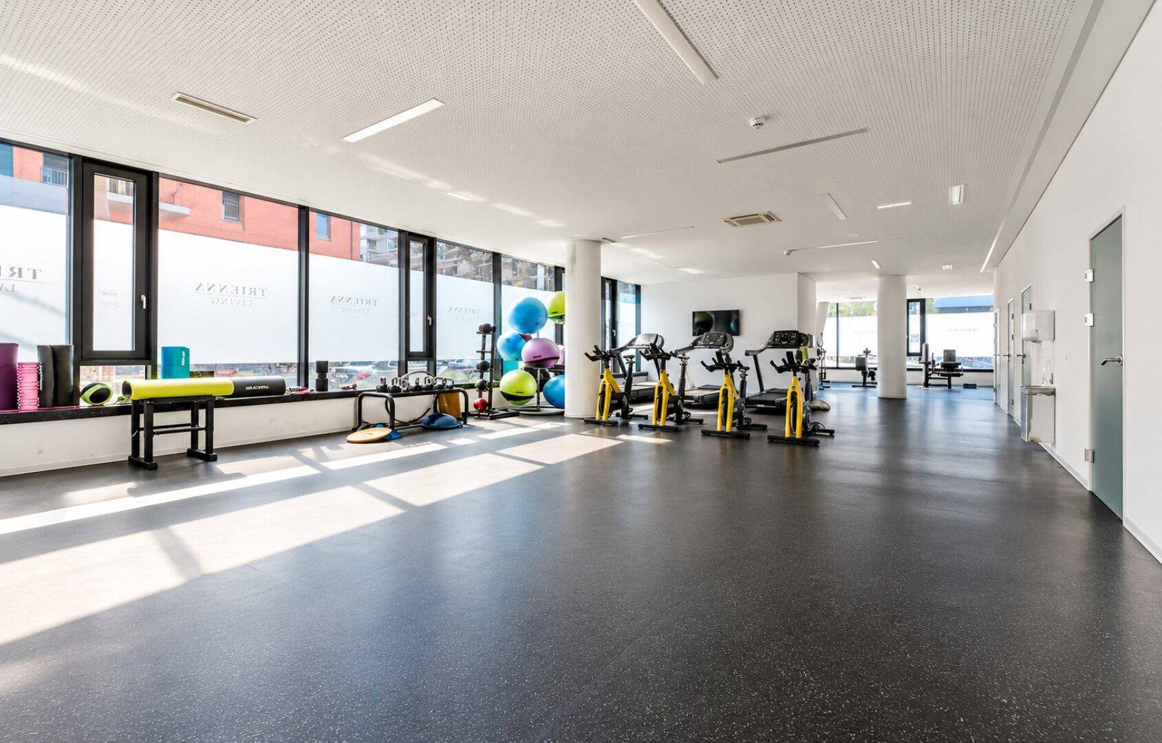 Geräumiger Fitnessraum mit verschiedenen Trainingsgeräten und großen Fenstern, ideal für Sport.