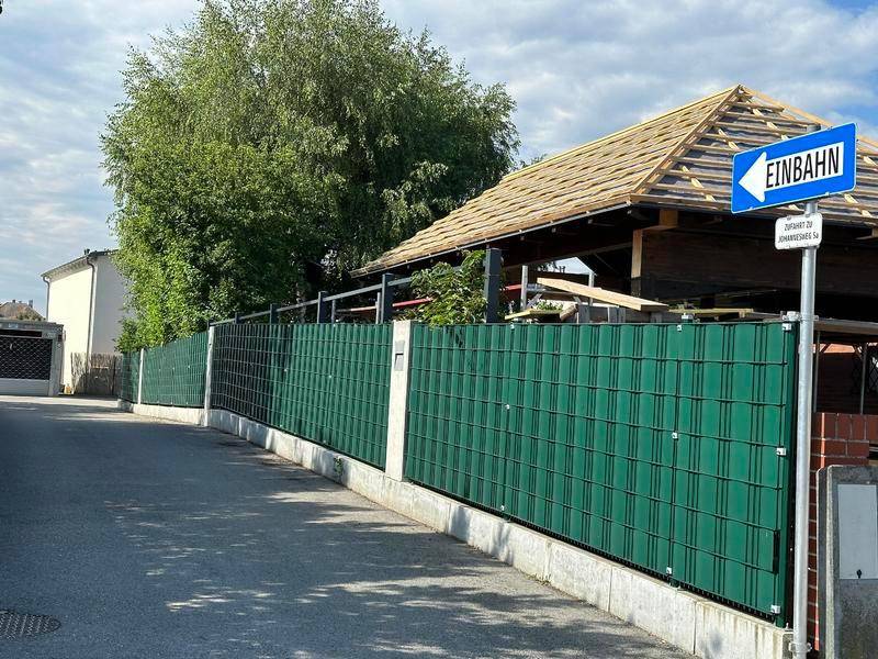 Grüner Zaun entlang einer Straße mit einem Gebäude im Bau und einem Einbahnstraßenschild.