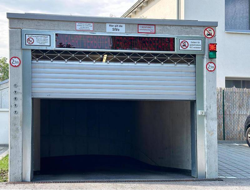 Einfahrt zu einer Tiefgarage mit Rolltor, Höhenbegrenzung und verschiedenen Warnschildern.