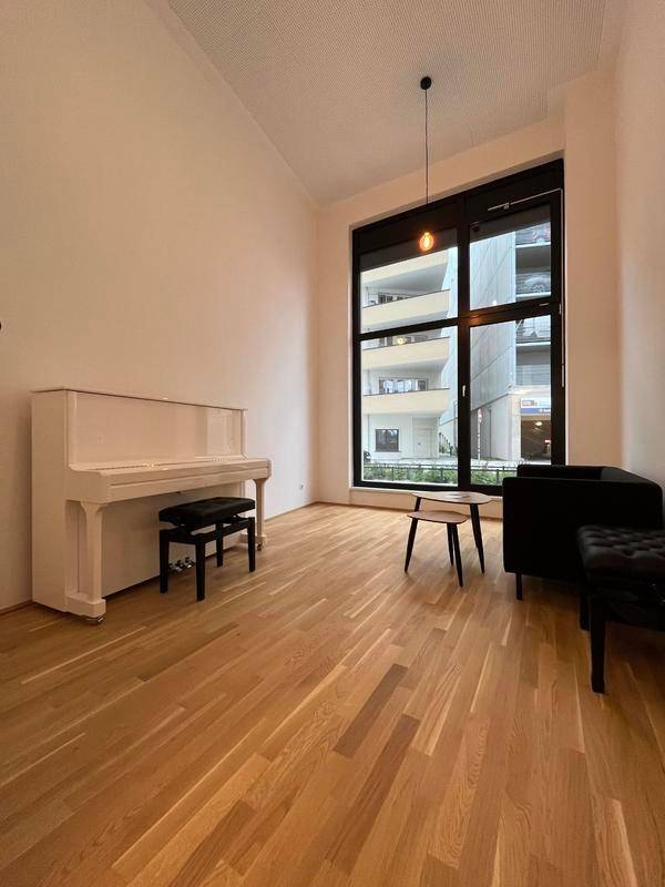 Geräumiges Wohnzimmer mit einem weißen Klavier, Holzboden und einem großen Fenster mit Blick auf andere Gebäude.