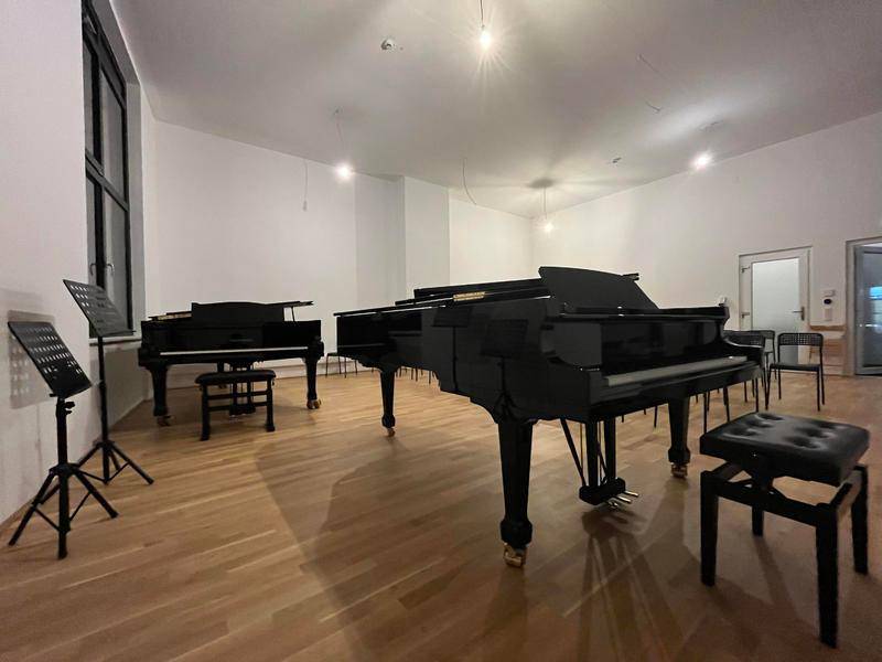 Nahaufnahme von zwei glänzenden schwarzen Flügeln in einem hellen Raum mit Holzfußboden und Musikständern.