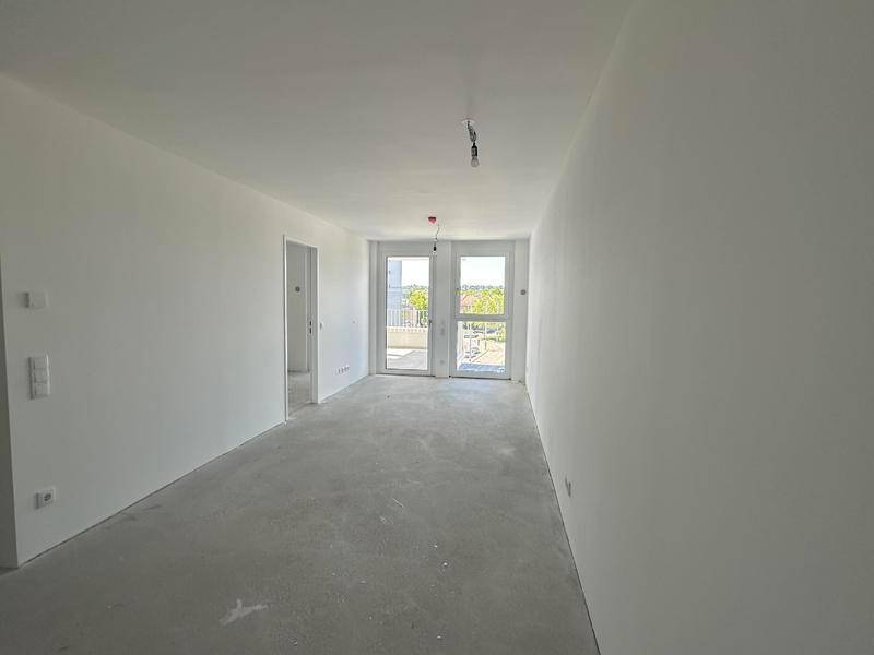 Geräumiger, unmöblierter Wohnbereich mit Rohbetonboden und großen Fenstern zum Balkon. Viel Tageslicht.