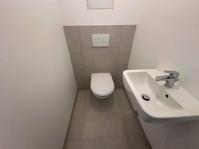 Separates WC mit wandhängender Toilette, kleinem Waschbecken und grauen Fliesen. Praktisch und platzsparend.