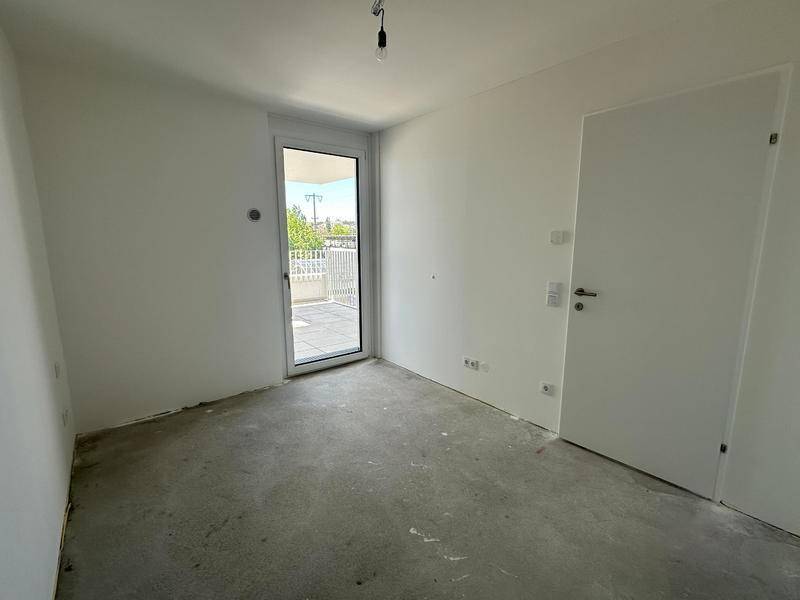Unmöblierter Raum mit Rohbetonboden und Zugang zu einer Terrasse, ideal als Schlafzimmer oder Büro. Hell und offen.