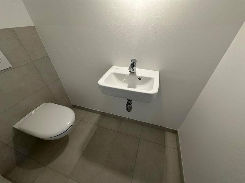 Kleines Gäste-WC mit hellgrauen Fliesen, einer Toilette und einem wandmontierten Waschbecken.