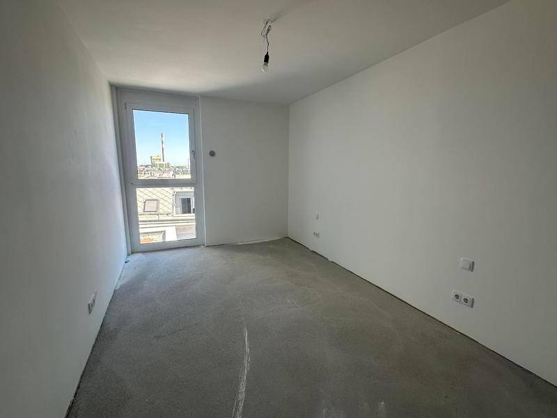 Unmöbliertes Zimmer mit weißen Wänden, Betonboden und großem Fenster mit Blick auf die Stadt.