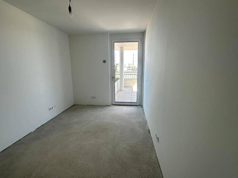 Unmöbliertes Schlafzimmer mit weiß gestrichenen Wänden, Betonboden und Zugang zu einem Balkon.