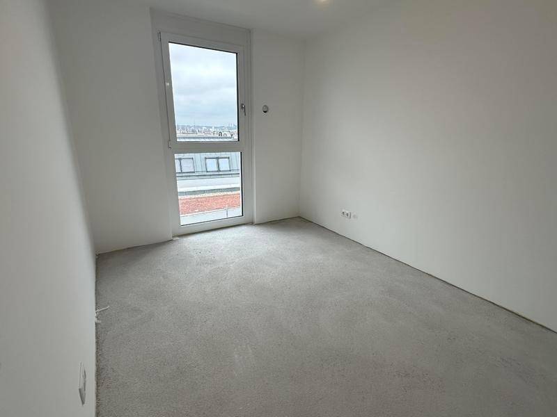 Unmöbliertes Schlafzimmer mit großem Fenster, Rohbetonboden und weißen Wänden, bietet viel Tageslicht.