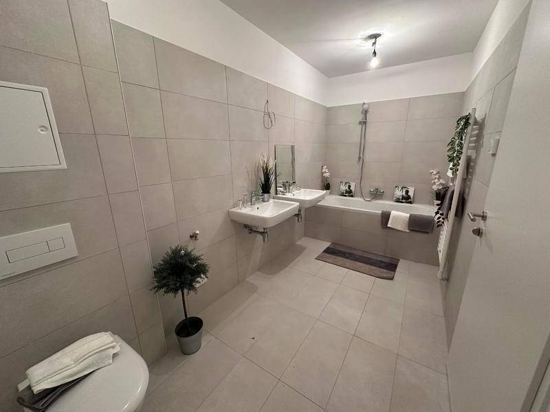 Modernes Badezimmer mit hellen Fliesen, Doppelwaschbecken und Badewanne mit Duschfunktion.