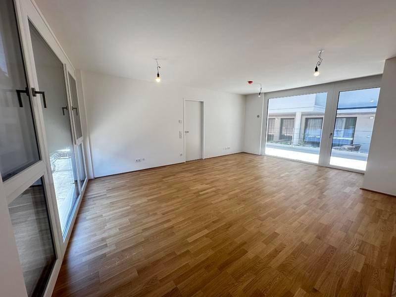 Geräumiges, leeres Wohnzimmer mit Holzboden und großen Fenstern, die viel Tageslicht hereinlassen.