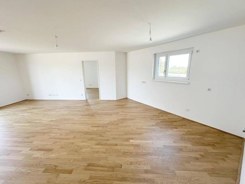 Geräumiger, leerer Wohnbereich mit Holzboden, weißen Wänden und einem großen Fenster.