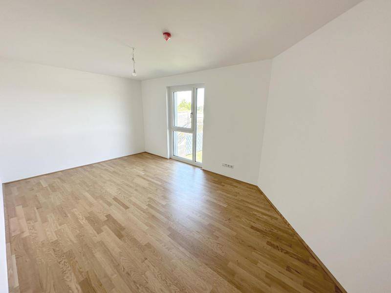 Helles, leeres Zimmer mit Holzboden und einer Glastür, die zu einem Außenbereich führt.