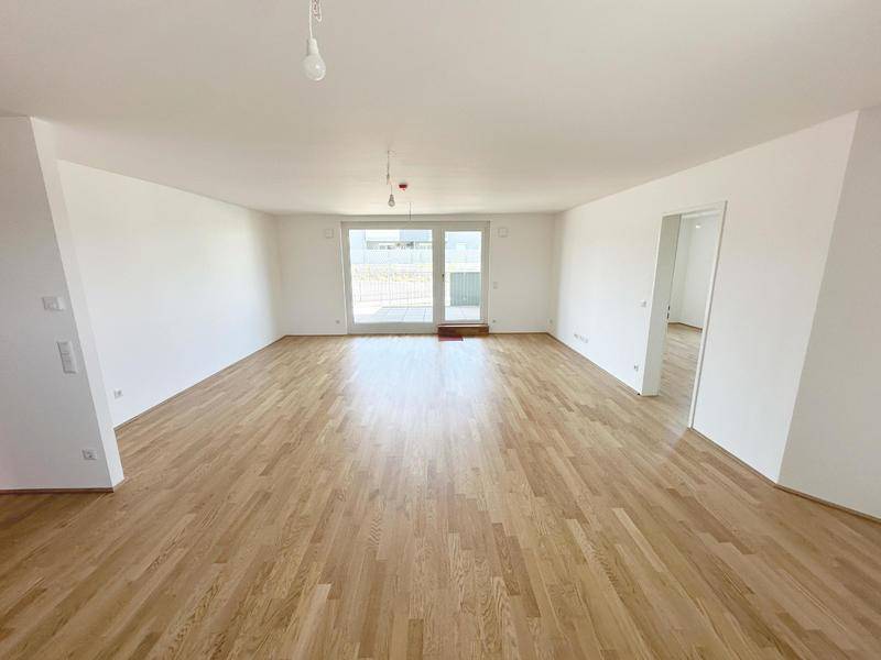 Heller, leerer Wohnbereich mit Holzboden und großem Fenster sowie Zugang zu einem Außenbereich.